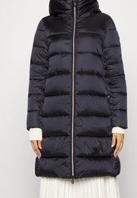 Pufjakke i mørk navy, med lynlås foran, tilpasset hætte og horisontal quiltet design. Blød, skinnende tekstur med ribbede manchetter.