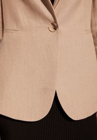 Beżowa marynarka z teksturowanego materiału, z zapięciem na jeden guzik i subtelnym łukiem u dołu, zestawiona z czarną prążkowaną odzieżą pod spodem.