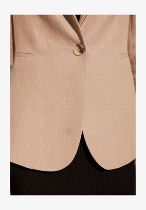 Beżowa marynarka z teksturowanego materiału, z zapięciem na jeden guzik i subtelnym łukiem u dołu, zestawiona z czarną prążkowaną odzieżą pod spodem.