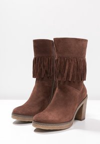 Stivaletti alla caviglia in camoscio marrone con dettagli a frange sulla parte superiore, punta quadrata e tacco impilato in legno. Cerniera laterale per una facile calzata.