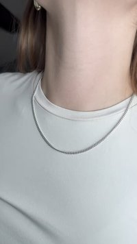 Stříbrný řetízek s lesklým, hladkým povrchem leží na světlé, dobře padnoucí košili. Na textilu košile je viditelná jemná textura.