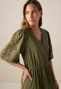 Robe vert olive avec un décolleté en V, des manches courtes bouffantes ornées de broderies florales, et une taille cintrée avec un cordon de serrage. Texture de tissu douce.