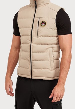 Beige quiltet vest med forreste lynlås og brystlomme, båret over en sort kortærmet skjorte på en stående mand.