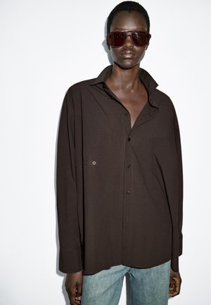 Bruine oversized button-up shirt met een kraag, voorzien van een borstzak, gedragen met lichtblauwe denim jeans en donkere zonnebril.