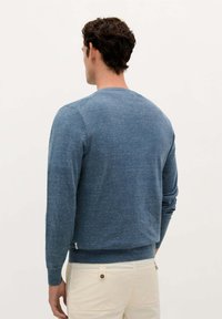 Blauwe gebreide trui met lange mouwen, ronde hals en geribbelde zoom. Stof heeft een gestructureerde heidelook. Draagt over lichtgekleurde broeken.