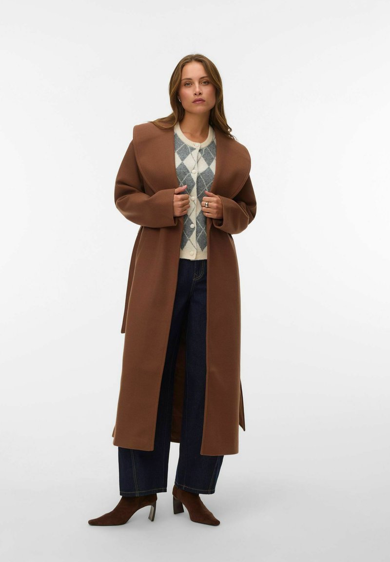 Manteau long marron avec larges revers, tissu texturé, devant ouvert, associé à un pull léger à motif losange et un jean foncé.