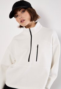 Hvid fleece pullover med høj krave, sort lynlås, brystlomme og forstærkede syninger; parret med en sort kasket.