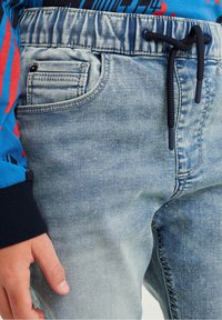 Joggers in denim azzurro chiaro con vita elastica, cordino, tasca laterale e texture sbiadita. Cuciture dettagliate attorno alla tasca.