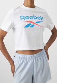 Bílý zkrácený tričko s modro-červeným logem Reebok. Spárováno se světle modrými šortkami s elastickým pasem.