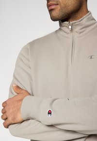 Ανοιχτό μπεζ μισό-φερμουαρ πουλόβερ κατασκευασμένο από απαλό ύφασμα. Διαθέτει λογότυπο στο στήθος και έγχρωμο μπαλώμα στο μανίκι. Ομαλή υφή σε όλο το ύφασμα.