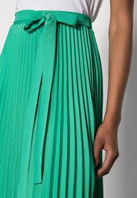 Gonna plissettata verde con cintura e dettaglio con legatura. Realizzata in un materiale liscio con un design strutturato e una leggera lucentezza.