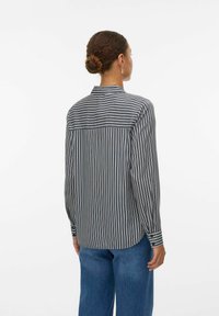 Kvinde med hår i en knold iført en sort og hvid lodret stribet skjorte og blå jeans, stående med ryggen mod kameraet foran en hvid baggrund.