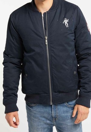 Mann trägt eine marineblaue Bomberjacke mit einem kleinen Astronauten-Graphic auf der linken Brust und eine blaue Jeans.