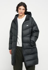 Abrigo acolchado negro con capucha, diseño acolchado y un sutil logotipo blanco de Nike en el pecho. La cremallera frontal se extiende hasta la mitad del muslo.