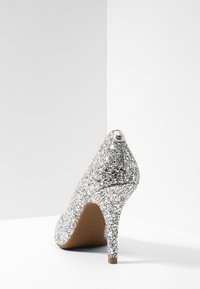 Escarpin à talon haut en paillettes argentées vu de l'arrière, avec une semelle marron et un petit détail en métal marqué au sommet du talon.