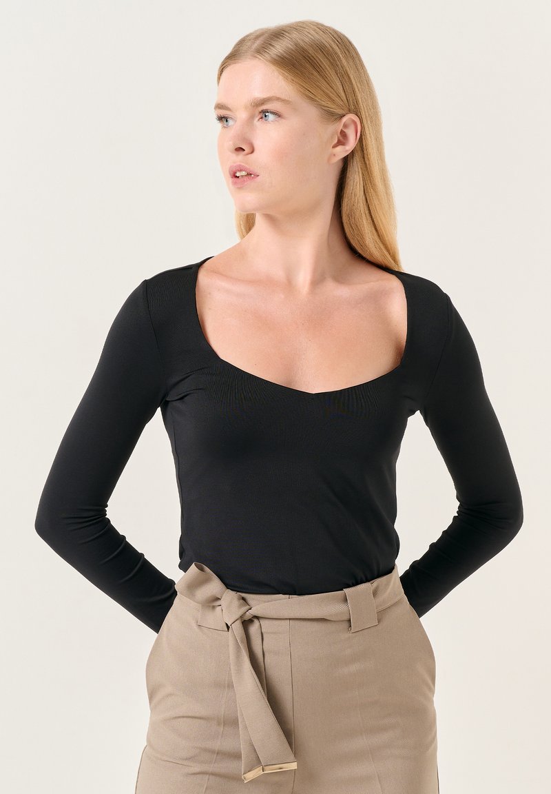 Top nero a maniche lunghe con scollo a barca, realizzato in un tessuto morbido. Indossato con pantaloni beige a vita alta, dotati di una cintura annodata.