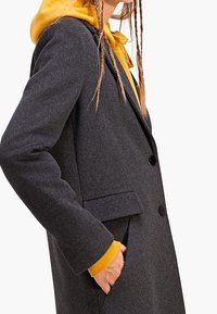 Abrigo gris de mezcla de lana con un corte entallado, que cuenta con dos botones negros, un bolsillo lateral y una capucha amarilla a contraste que asoma por debajo.