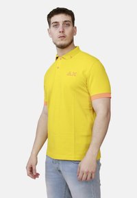 Uomo che indossa una polo gialla brillante con bordi arancioni e jeans azzurri chiari, in piedi e che guarda leggermente di lato.