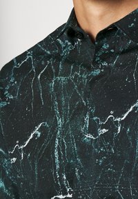 Camicia nera con un motivo marmorizzato con accenti teal e bianchi. Collo button-down, tessuto testurizzato e bottoni neri piccoli.