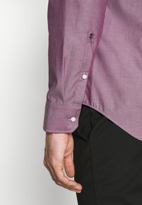 Pravá ruka osoby ve fialové košili na knoflíky, s manžetou a černými kalhotami, uvolněná vedle těla, na rukávu malá vyšívaná květina.