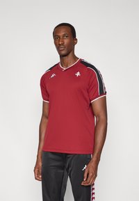 Μπορντό αθλητική μπλούζα με λευκές λεπτομέρειες, σχέδιο με V λαιμόκοψη και μαύρες πανελ στους ώμους. Το ύφασμα φαίνεται λείο με κοντά μανίκια. Φαίνεται το λογότυπο της Kappa.