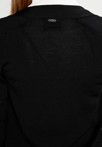 Schwarzer Strickpullover mit strukturiertem Finish. Mit Rundhalsausschnitt und kleinem silbernen Logo auf der Rückseite, nahe dem Kragen.