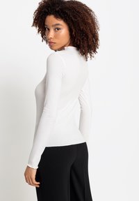 Witte langmouwige fitted top met geribbelde textuur en hoge halslijn, gecombineerd met zwarte wijde broek, bekeken vanaf de achterkant.