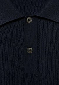 Polo de color azul marino con una tela texturizada, que presenta un collar y una tapeta de dos botones en metal oscuro con un diseño de cruz.