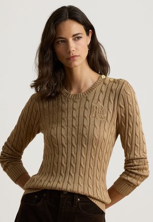 Femme aux cheveux foncés portant un pull en maille torsadée beige avec des boutons sur une épaule, debout avec les bras légèrement pliés.