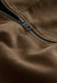 Nahaufnahme einer braunen Wildlederjacke mit einem teilweise zippten silbernen Metallreißverschluss und sichtbaren Nähdetails.