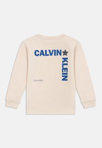 Béžové tričko s dlouhým rukávem s výrazným modrým nápisem "CALVIN KLEIN" a grafikou hvězdy. Měkká tkanina s uvolněným střihem.