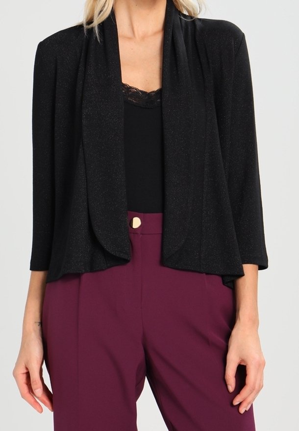 Femme portant un cardigan noir ouvert devant avec manches 3/4 sur un haut noir bordé de dentelle et un pantalon bordeaux taille haute avec un bouton doré.
