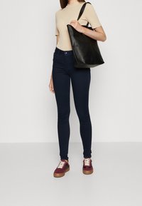 Tajta jeans i mörkblå denim, kombinerat med en beige åtsittande topp och vinröda sneakers. En svart läderhandväska bärs på ena axeln.