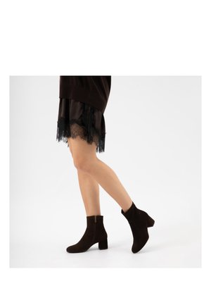 Beine einer Person, die einen schwarzen, mit Spitze besetzten Rock und schwarze Wildleder-Stiefeletten mit Blockabsätzen trägt, die auf einem weißen Hintergrund gehen.