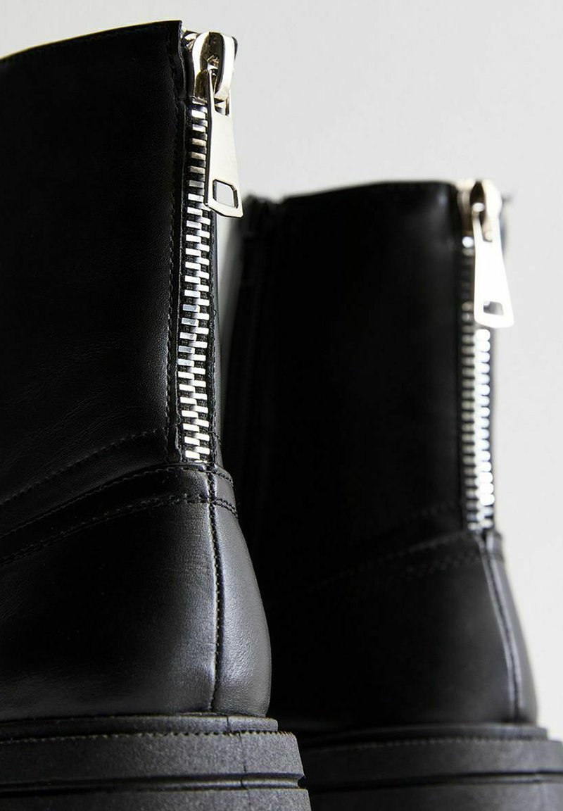 Stivaletti neri in pelle con cerniere argentate, caratterizzati da una suola spessa e dettagli cuciti. Le cerniere si estendono lungo i lati per facilitare l'indossamento.