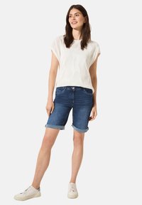 Haut tricoté blanc à manches courtes, associé à un short en jean bleu foncé à revers. Le mannequin porte des baskets blanches, se tenant devant un fond uni.