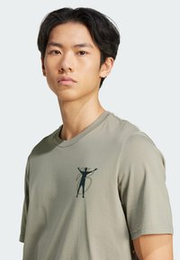 Olive green cotton t-shirt with a crew neck, featuring a dark silhouette graphic on the left chest and short sleeves.