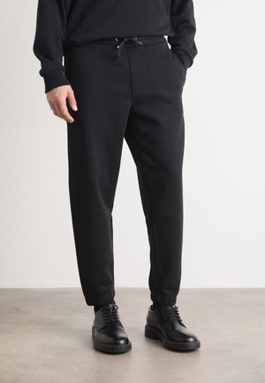 Mann trägt schwarze, zulaufende Jogginghosen mit Kordelzug in der Taille und schwarze Leder-Schnürschuhe und steht auf einem grauen Boden vor einer schlichten Wand.
