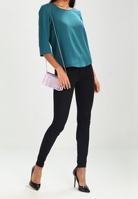 Donna che indossa una maglietta teal a tre quarti, pantaloni skinny neri, tacchi alti neri e tiene in mano una piccola borsa a tracolla in tessuto leggero di colore viola chiaro.