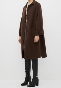 Femme portant un manteau en laine marron boutonné sur une robe à motifs avec des bottes noires hautes à talons, debout devant un fond uni.