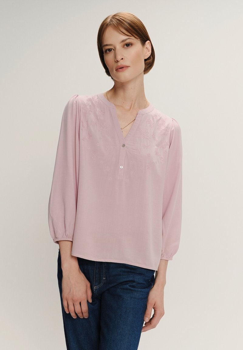 Donna con capelli corti castani che indossa una blusa rosa chiaro con ricami e maniche a tre quarti, abbinata a jeans blu scuro, in piedi contro uno sfondo semplice.