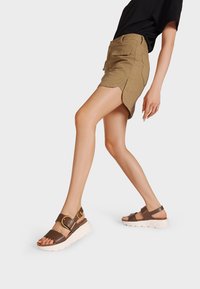 Sandali platform marroni con suola bianca spessa, caratterizzati da due cinghie marroni e un dettaglio con fibbia dorata. Abbinate a una gonna khaki.