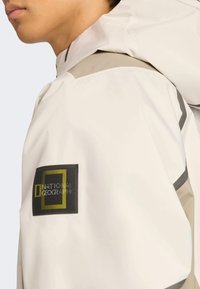 Hvid vandtæt jakke med en beige accent på skulderen, med et sort rektangulært patch, der viser National Geographic-logoet.