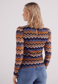 Strak zittend langemouwen top in een meerkleurig zigzagpatroon met oranje, blauw en crème tinten; sleutelgatdetail aan de achterkant en textuurstof.