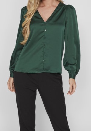Nainen, joka pitää päällään tummanvihreää satiinista V-kaulaista paitapuseroa, jossa on pitkät pönkköhihat, ja mustat housut, seisoo vaaleaa taustaa vasten.