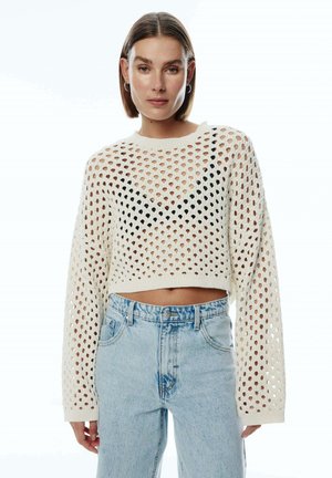 Cremefarbenes, kurz geschnittenes Sweatshirt mit großem offenen Strickmuster, langen Ärmeln und rundem Ausschnitt, kombiniert mit hellblauen, hochtaillierten Jeans.