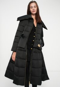 Long black puffer coat with a wide collar, padded sections, and gold zipper. Features a front pocket with a branded detail and button closure.