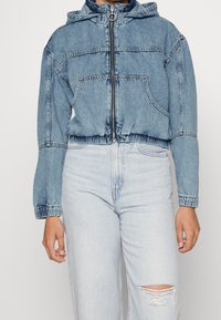 Džínová bunda s kapucí, světle modrý odstín, přední zip, elastický lem, dlouhé rukávy, dvě přední kapsy a kontrastní šití.