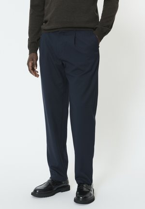 Marineblå bukser med en afslappet pasform, der har frontfolder, sidelommer og glat stof, kombineret med sorte sko og en mørk sweater.