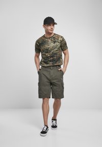 T-shirt verde mimetico abbinato a pantaloni cargo verdi oliva con tasche laterali. Completa il look con un cappellino nero e sneakers nere.
